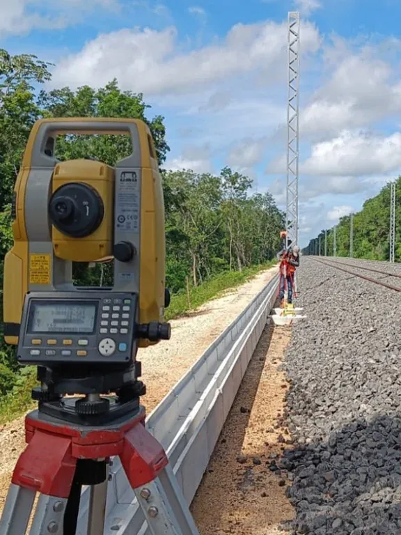 Estación total sobre tripié junto a la vía, con postes de catenaria al fondo